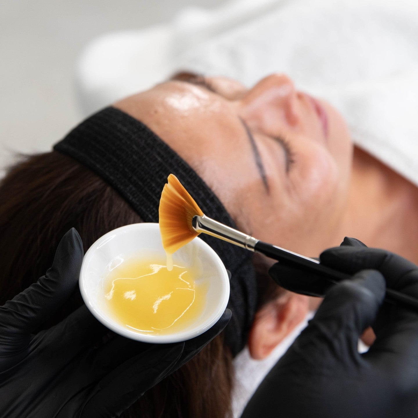 Person receiving a facial treatment with a brush and bowl of yellow liquid.