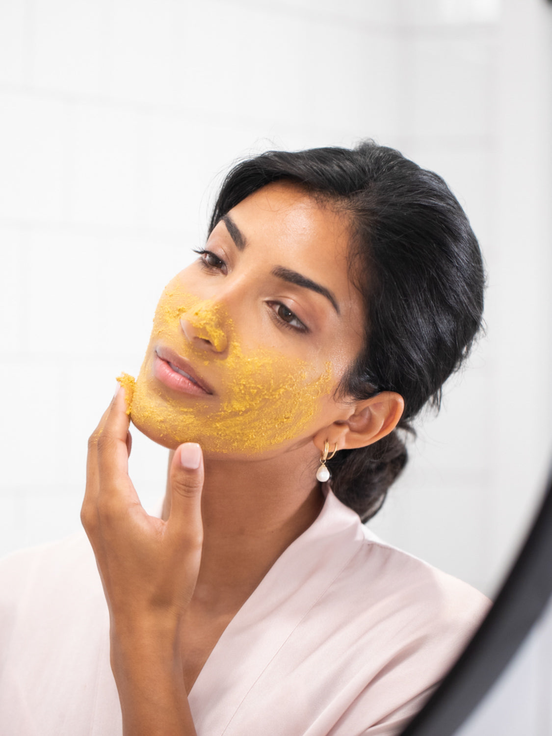 Woman applying a yellow facial mask in front of a mirror.