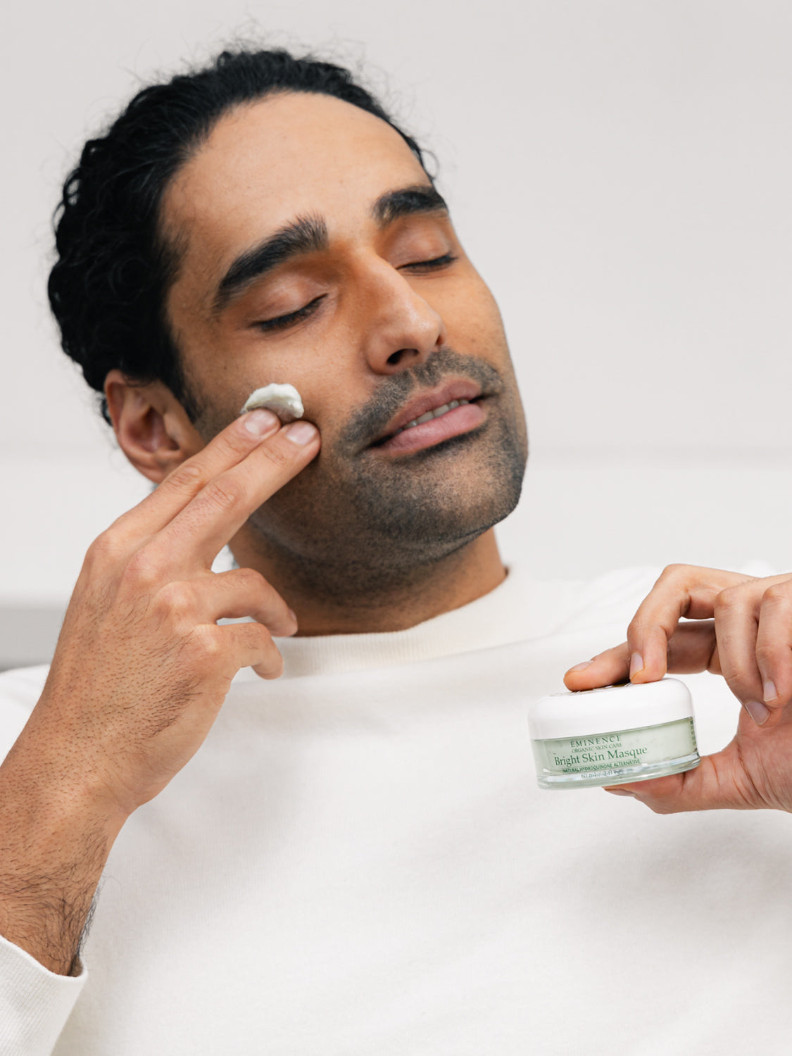 Man applying cream to his face with a jar of cream in his other hand against a white background