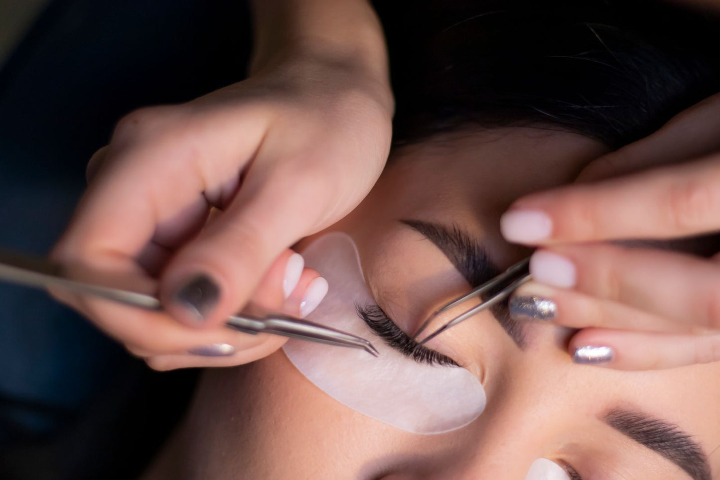 Close-up lash work with fine tweezers separating the lashes over a white under-eye pad.