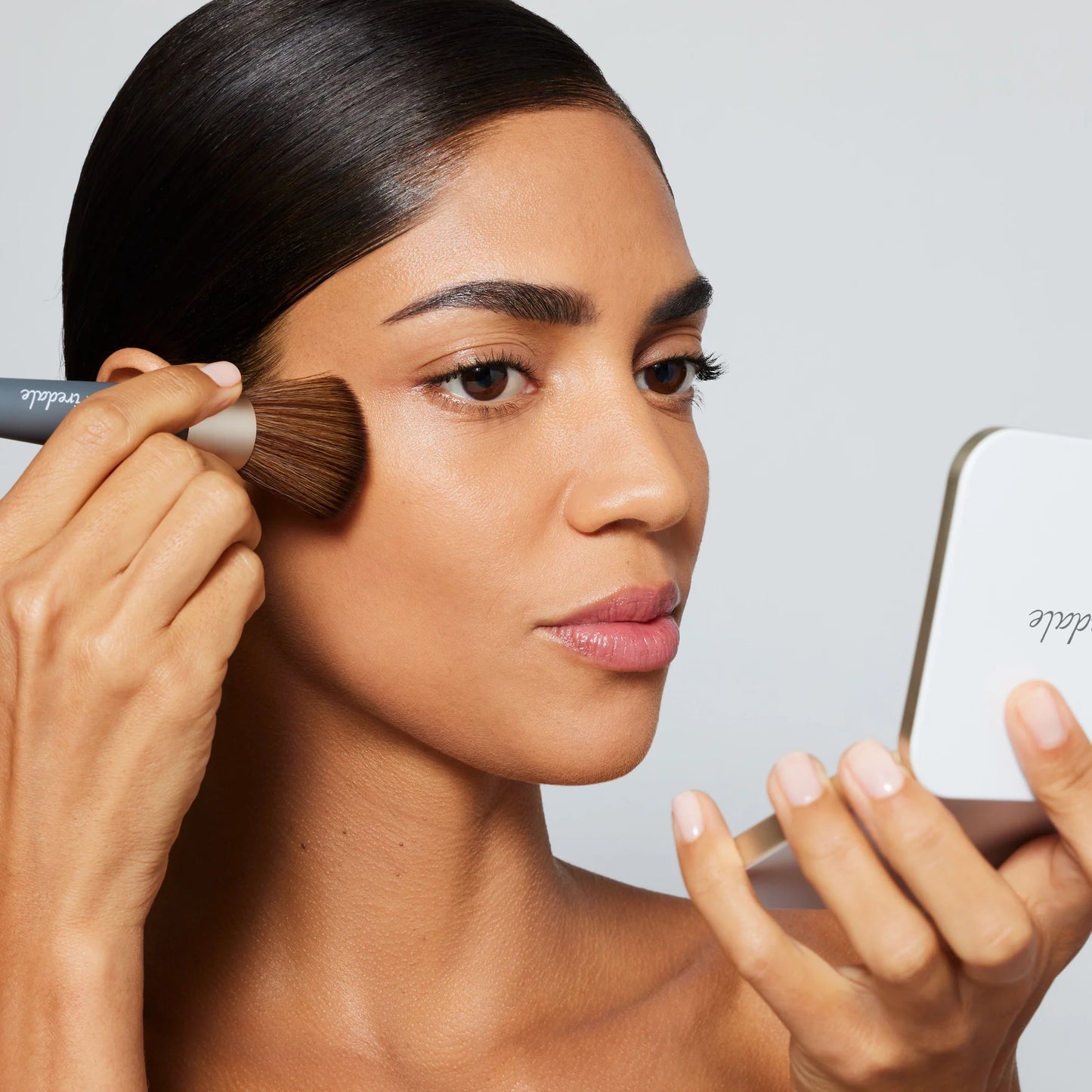 Woman applying makeup with a brush and mirror on a plain background