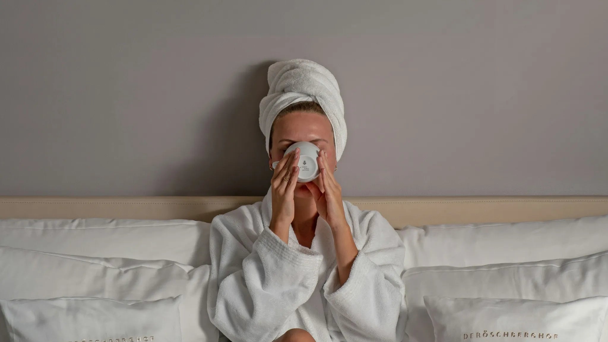 Person in a white robe and towel on head applying cream to face in a bed with white bedding.