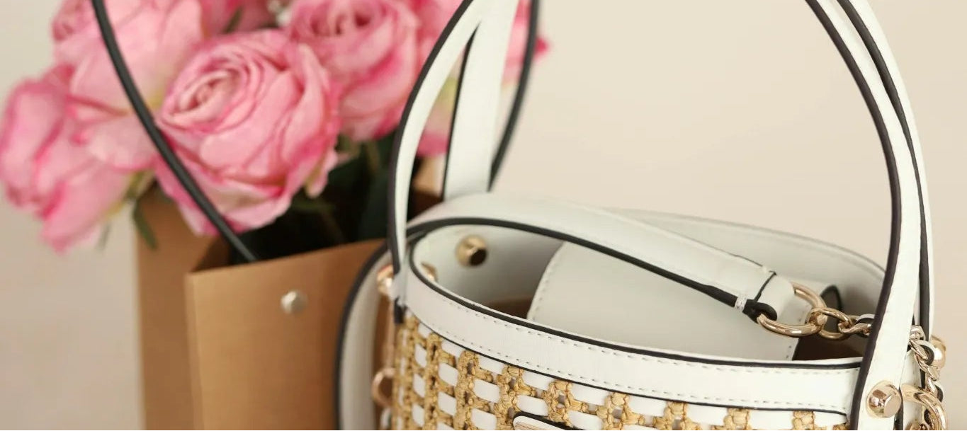 Woven handbag with a chain detail next to pink flowers in a box on a light surface.
