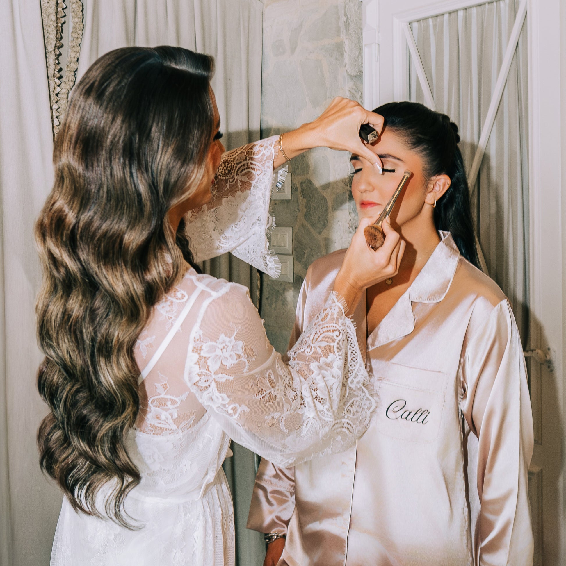 Woman getting makeup applied by another woman in a bedroom setting.