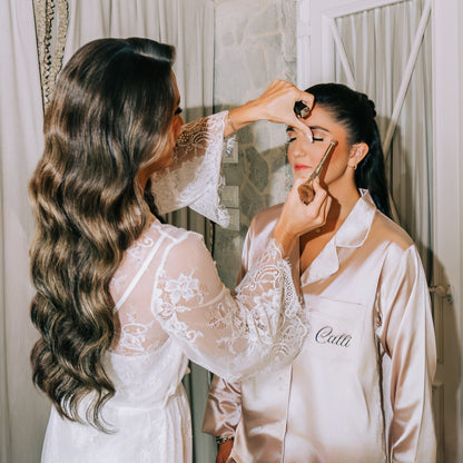 Woman getting makeup applied by another woman in a bedroom setting.