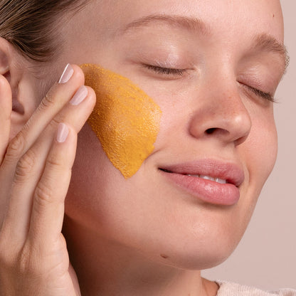 Woman applying a yellow facial mask to her cheek with a finger.