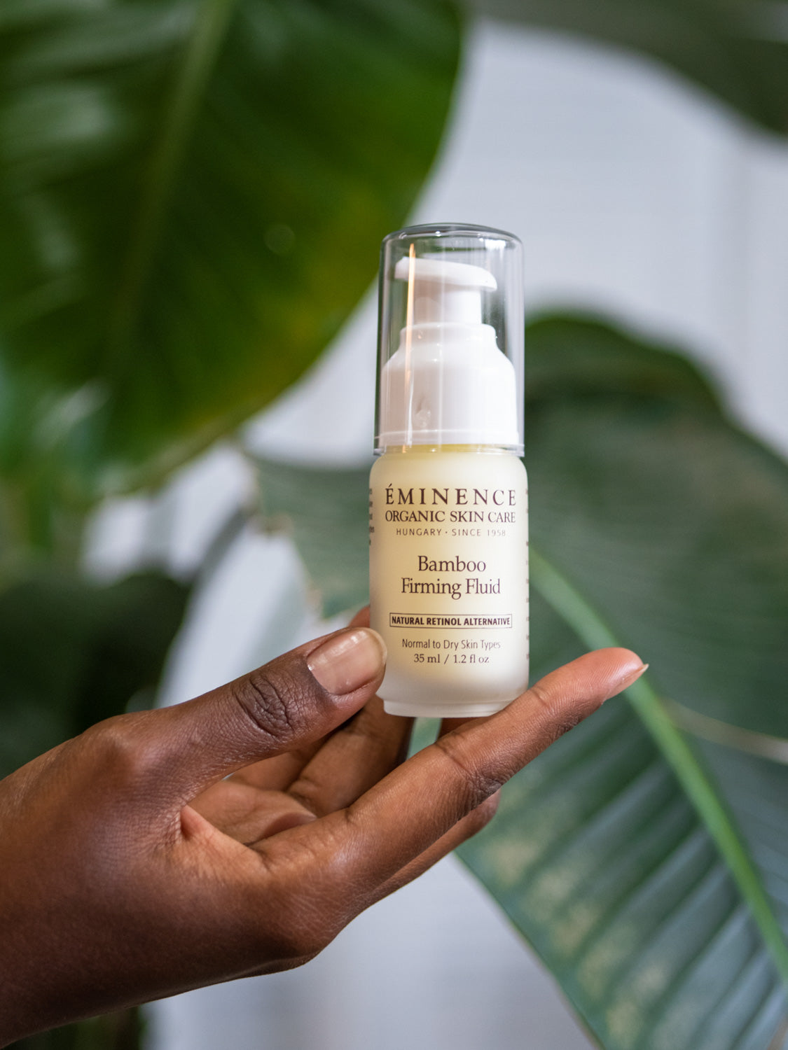 Hand holding a bottle of Eminence Bamboo Firming Fluid with green leaves in the background