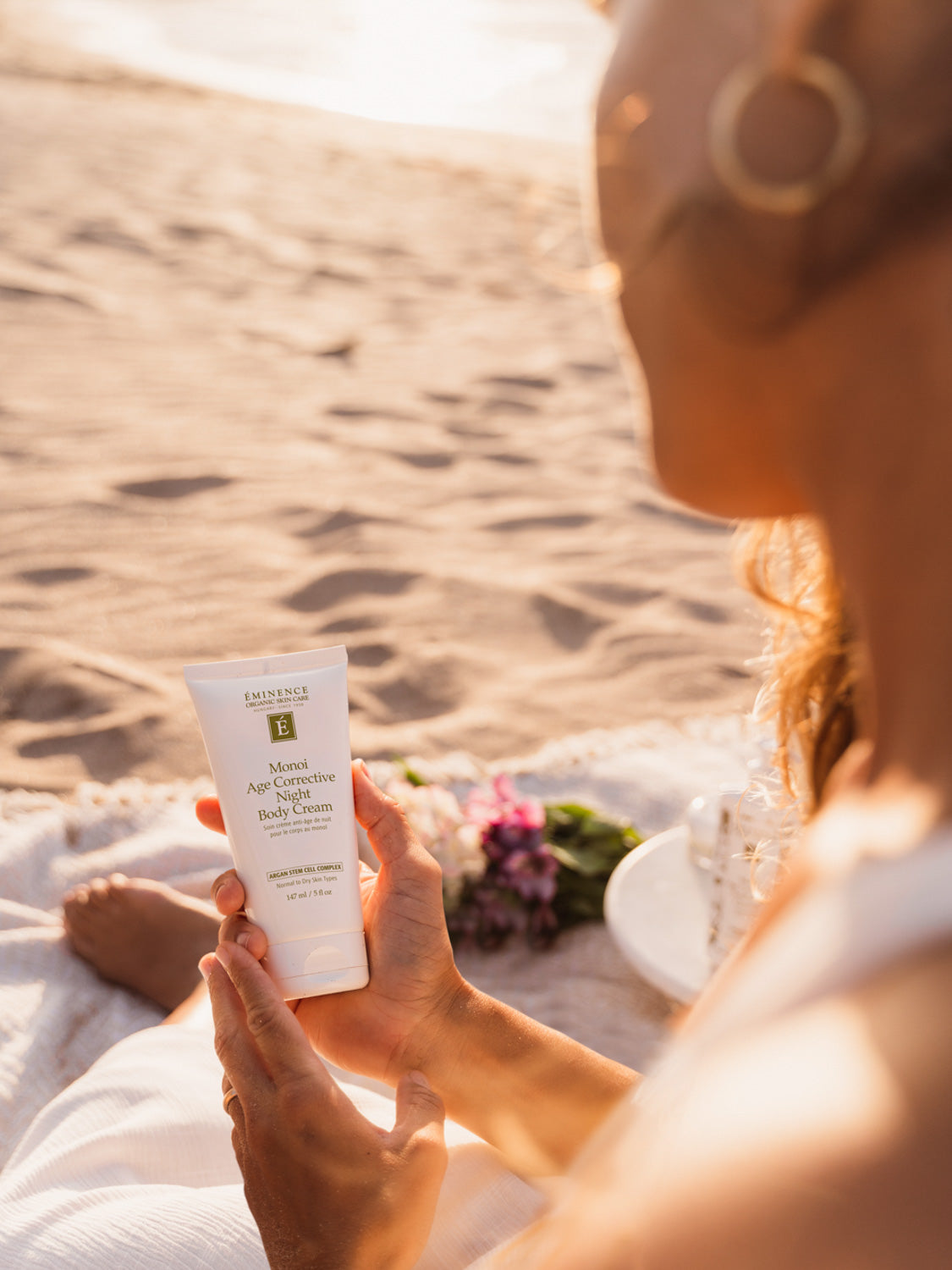 Person holding a skincare product on a beach with sand and sun in the background