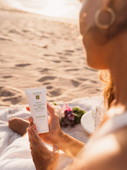 Person holding a skincare product on a beach with sand and sun in the background