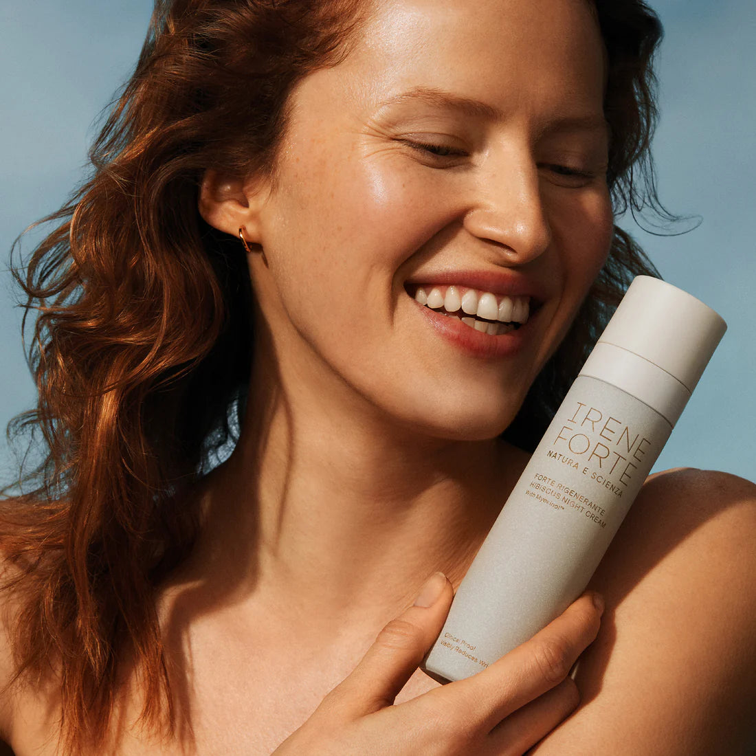 Woman holding a bottle of Irene Forte skincare product against a clear blue sky.