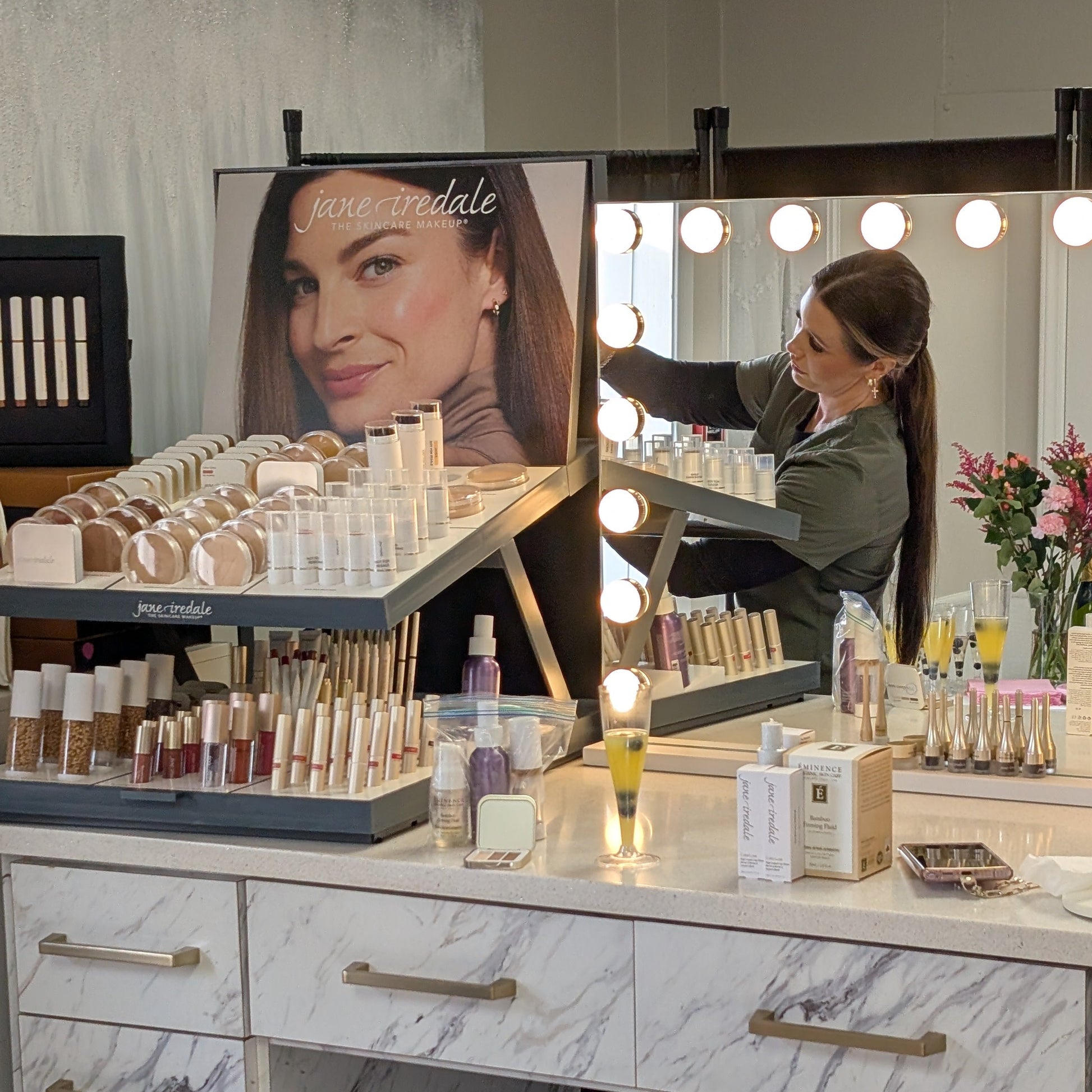 Makeup counter with various products and a woman applying makeup in a well-lit room.