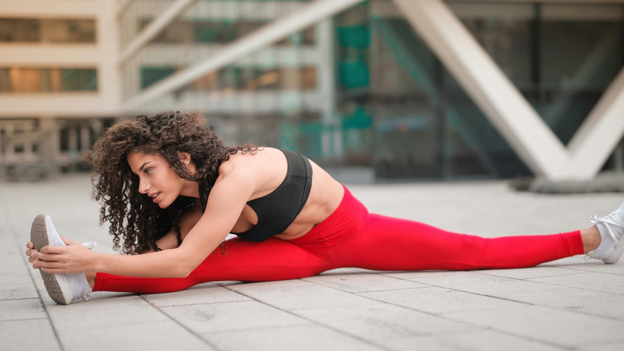 Woman in red leggings and black top stretching outdoors with modern building in background