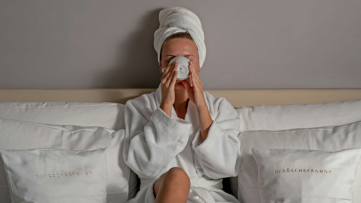 Person in a white robe and towel on head, sitting on a bed with white bedding.