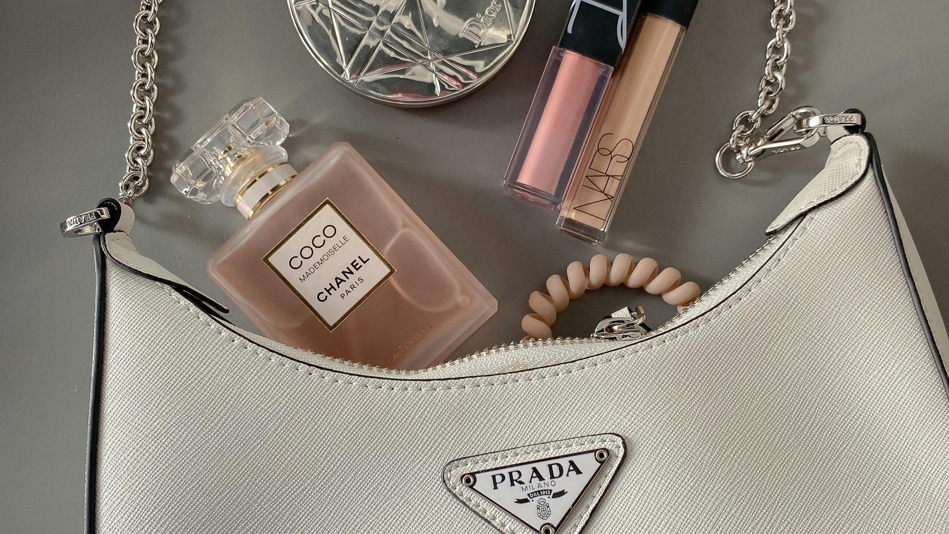 Prada handbag with makeup items including a perfume bottle and lip products on a neutral background