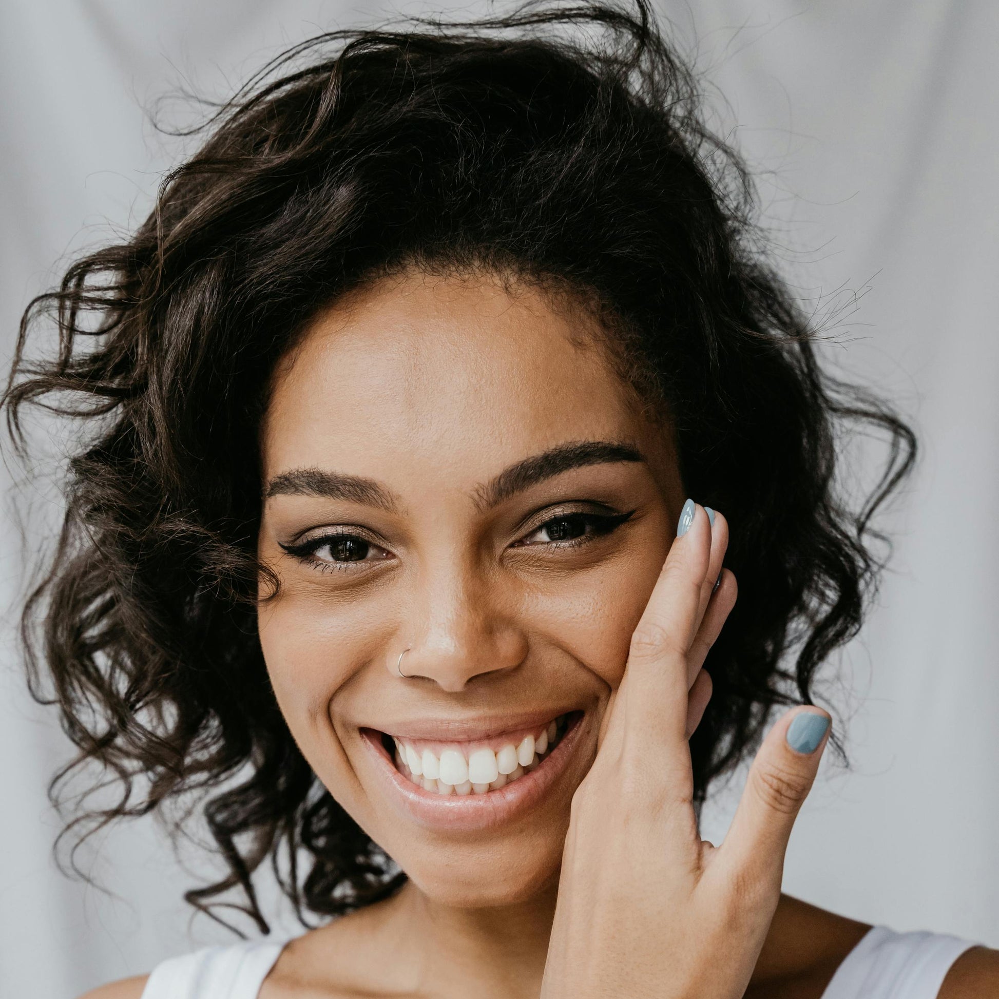 Woman with curly hair smiling and touching her face against a plain background