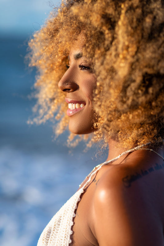Sunlit profile of a smiling woman with curly hair, sea and bright sky behind her.