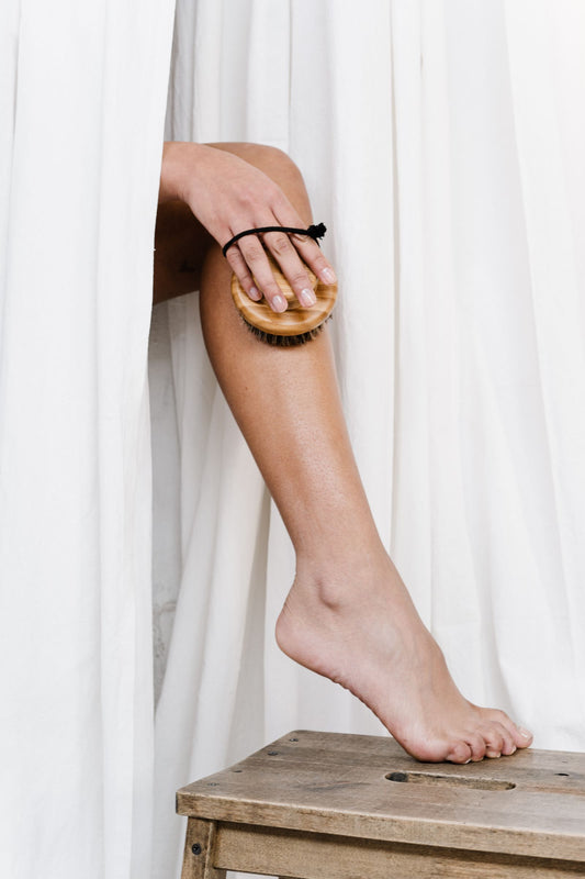 Lower leg exfoliation with a natural bristle body brush, set against soft white draping.