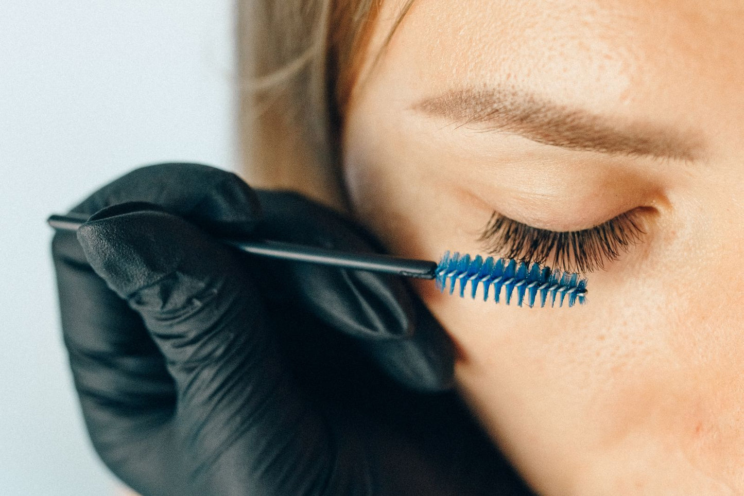 Lash tint being combed through closed lashes with a spoolie brush held in a gloved hand.
