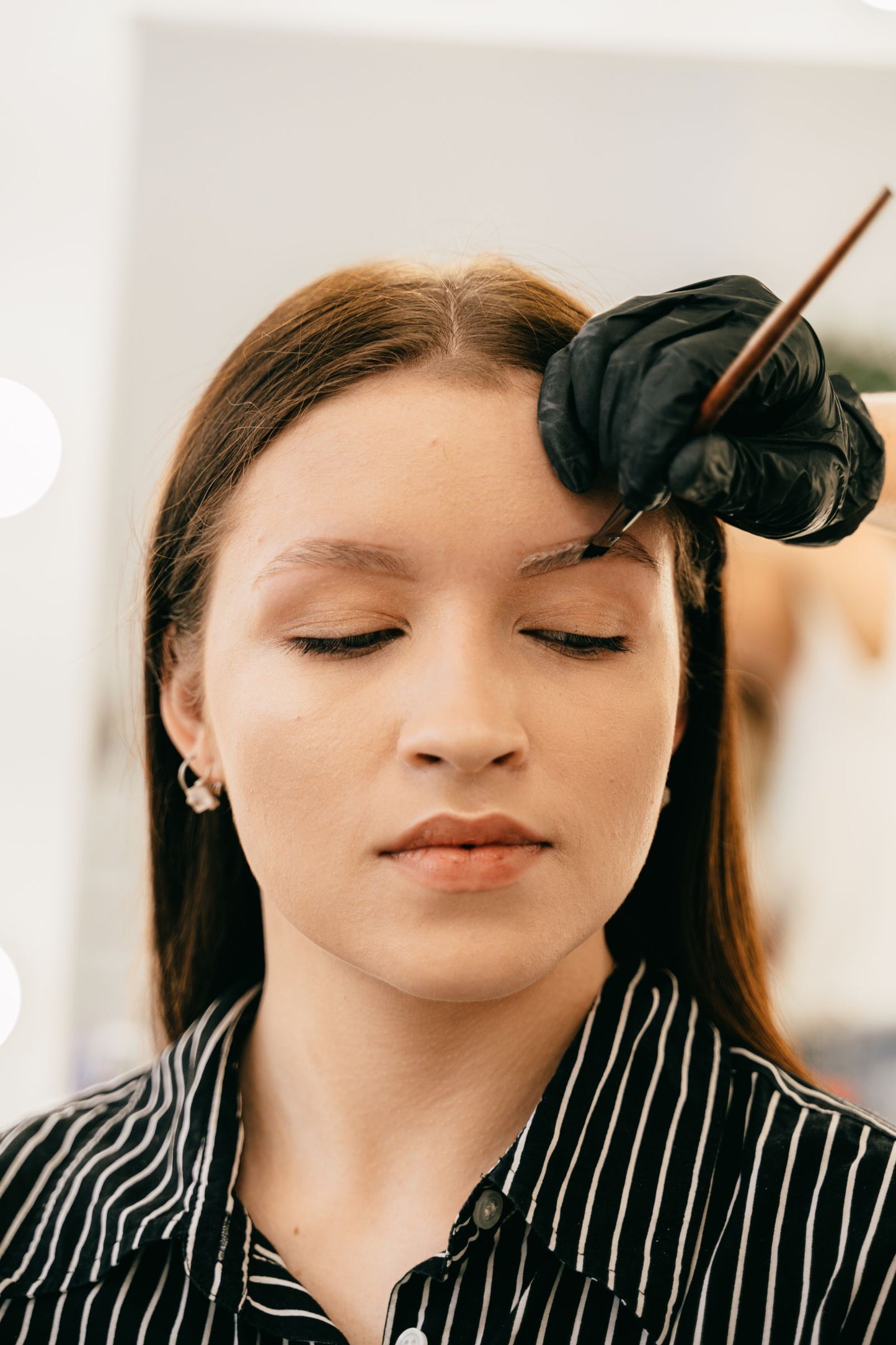 Brow tint being brushed onto the brow with a fine angled brush while the client's eyes stay closed.
