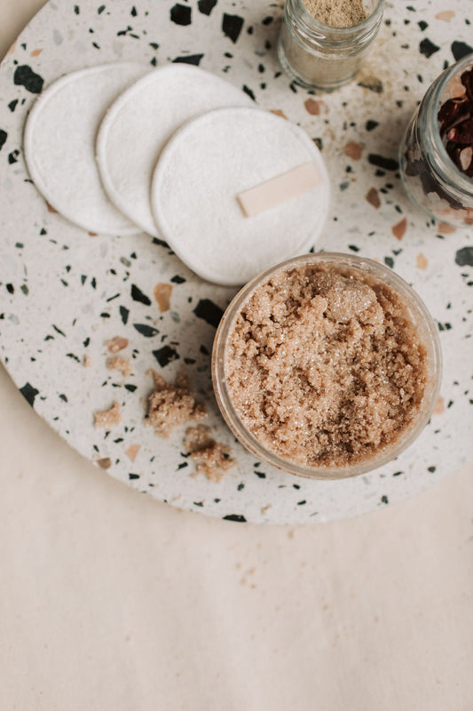 Brown sugar body scrub in a glass bowl on a terrazzo tray, with soft cotton rounds alongside.